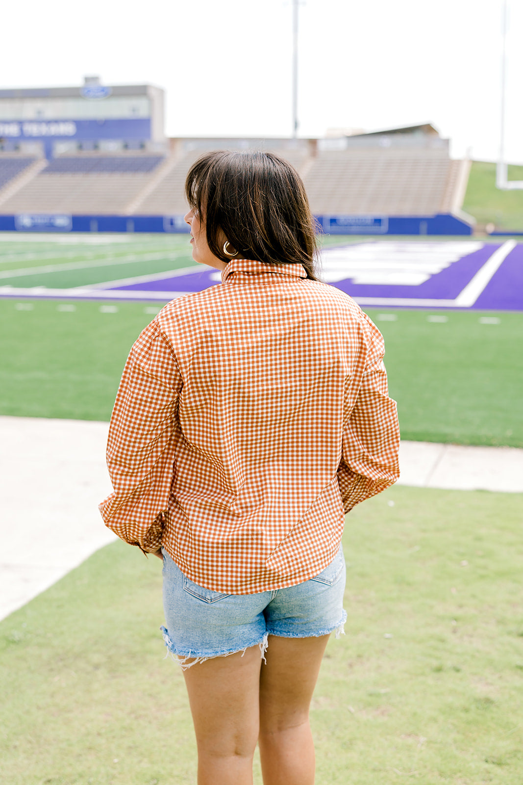 Woman with dark hair wearing a burnt orange gingham long-sleeve top, and light-wash denim shorts.