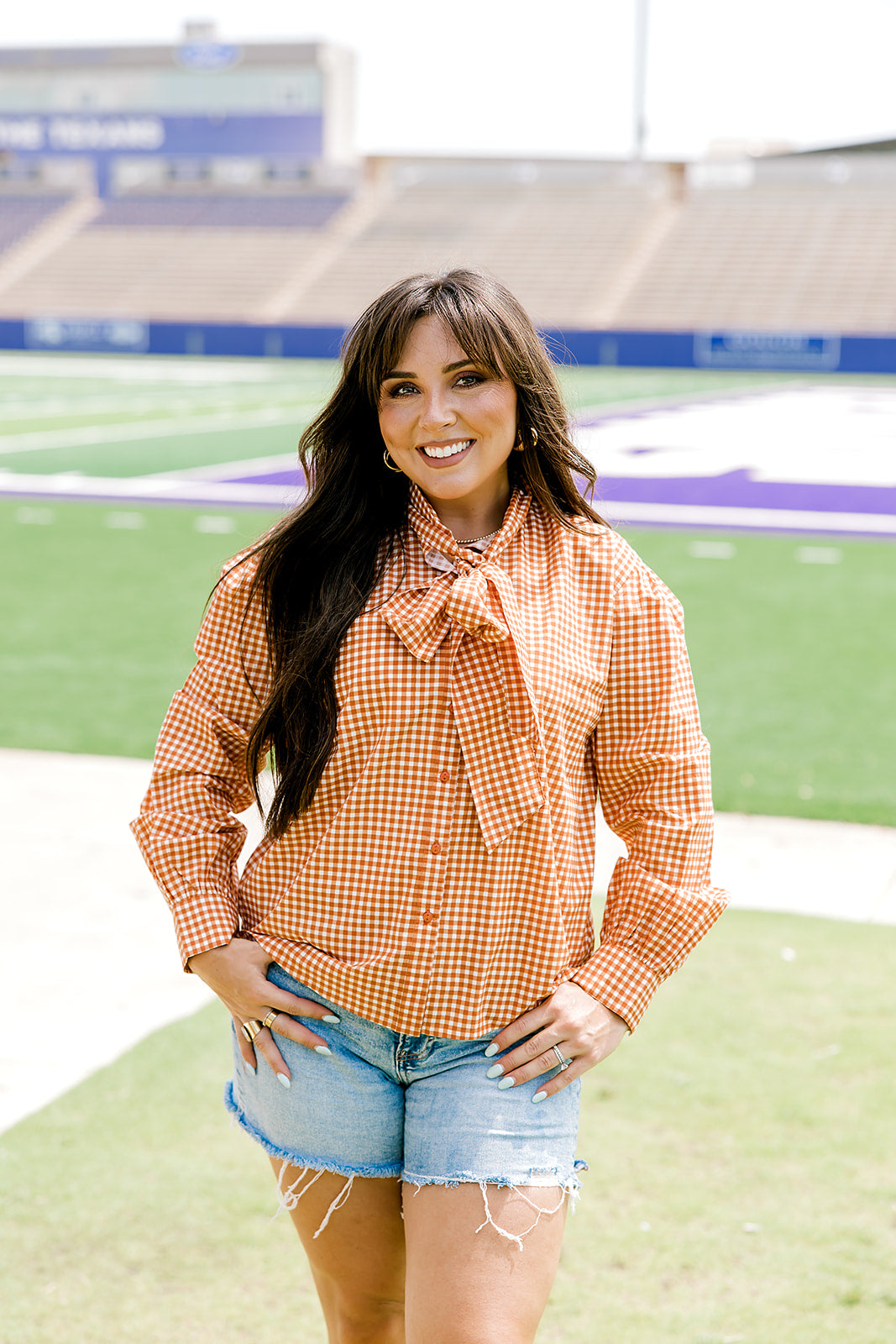 Woman with dark hair wearing a burnt orange gingham long-sleeve top, and light-wash denim shorts.