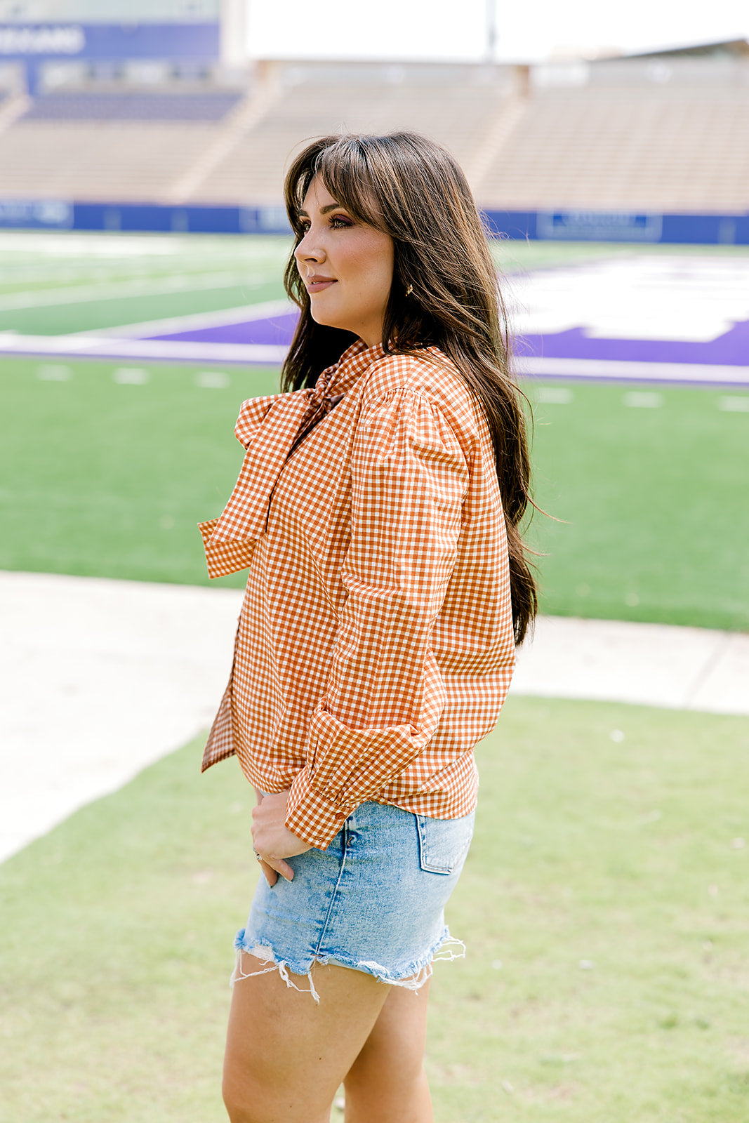 Woman with dark hair wearing a burnt orange gingham long-sleeve top, and light-wash denim shorts.