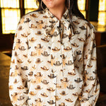Woman with dark hair wearing a long-sleeve button-down with multi-colored cowboy hats and tying feature in front of top. Model paired this top with light-wash denim jeans.