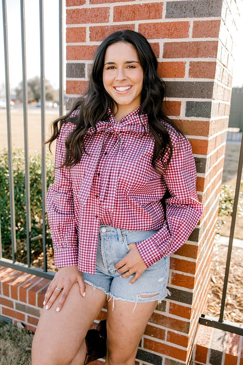 Woman with dark hair wearing a button down, maroon gingham long-sleeve with a bow tied in front, and light-wash denim shorts.