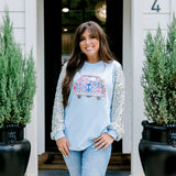 Woman with dark hair wearing a baby blue sweatshirt with iridescent sparkle sleeves; front of top has holiday truck embroidered on it. Model paired this top with light-wash denim jeans.