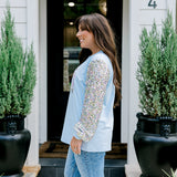 Woman with dark hair wearing a baby blue sweatshirt with iridescent sparkle sleeves; front of top has holiday truck embroidered on it. Model paired this top with light-wash denim jeans.
