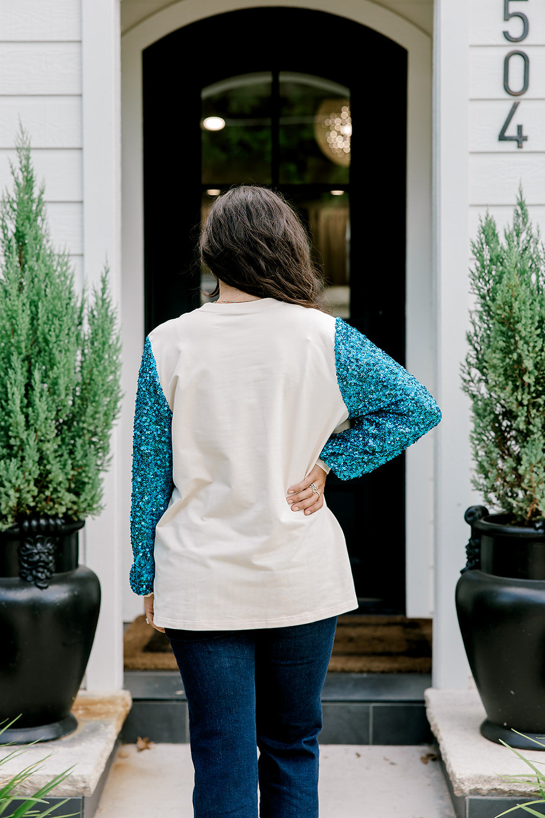 Woman with dark hair wearing a sweatshirt with turquoise sparkle sleeves & embroidery on front that says "Jesus Is The Reason". Model paired this top with dark-wash denim jeans.