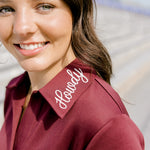 Woman with dark hair wearing a maroon dress with a close-up on howdy embroidery.