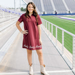 Woman with dark hair wearing a maroon midi dress with white embroidery along bottom, and cream colored boots.