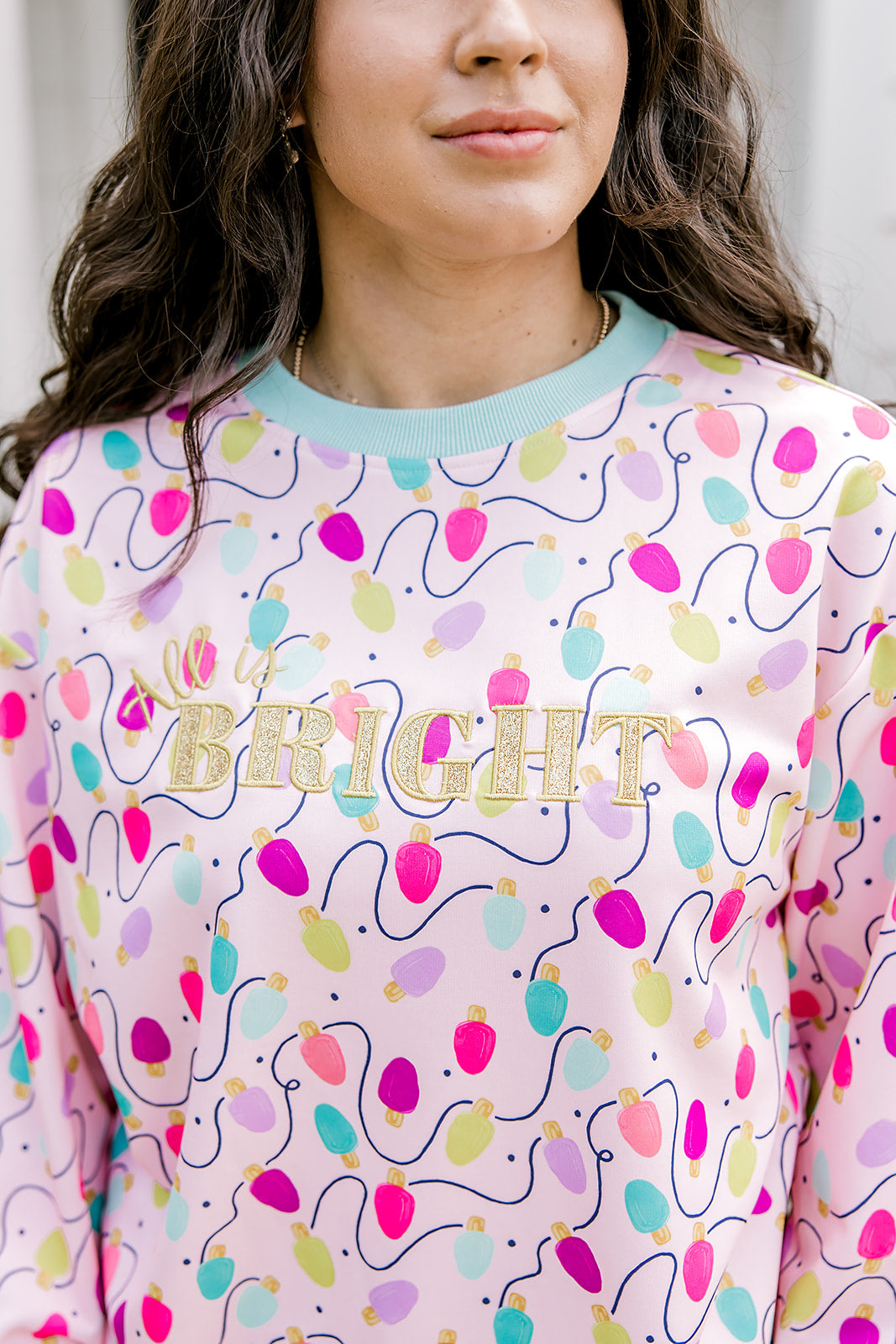 Woman with dark hair wearing a sweatshirt with Christmas light graphics  that are multi-colored. Model paired this top with dark-wash denim jeans.