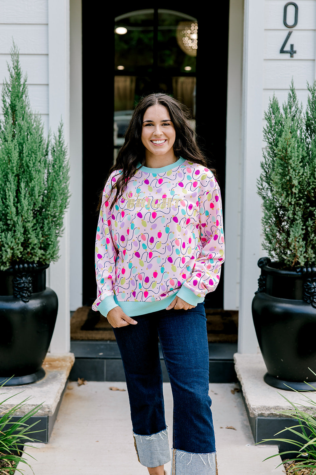 Woman with dark hair wearing a sweatshirt with Christmas light graphics  that are multi-colored. Model paired this top with dark-wash denim jeans.