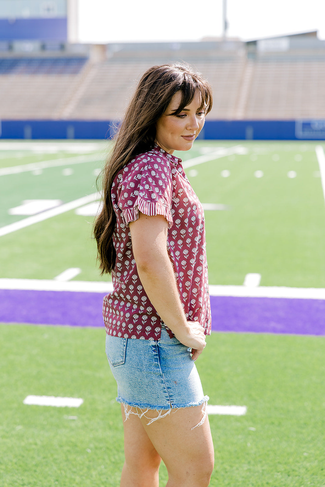 Woman with dark hair standing in a side profile position wearing the Stevie Top in a lifestyle scene. The focal point of this image is the striped ruffle sleeves as well as length of shirt and flower graphics. The model paired this tip with light-wash denim shorts.