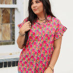 Woman with dark hair standing in a front profile position wearing the Stevie Top against a white background. The focal point of this image is the pink and orange floral, buttons down the front, as well as the low collar and ruffled sleeves. The model paired this top with dark-wash denim jeans.