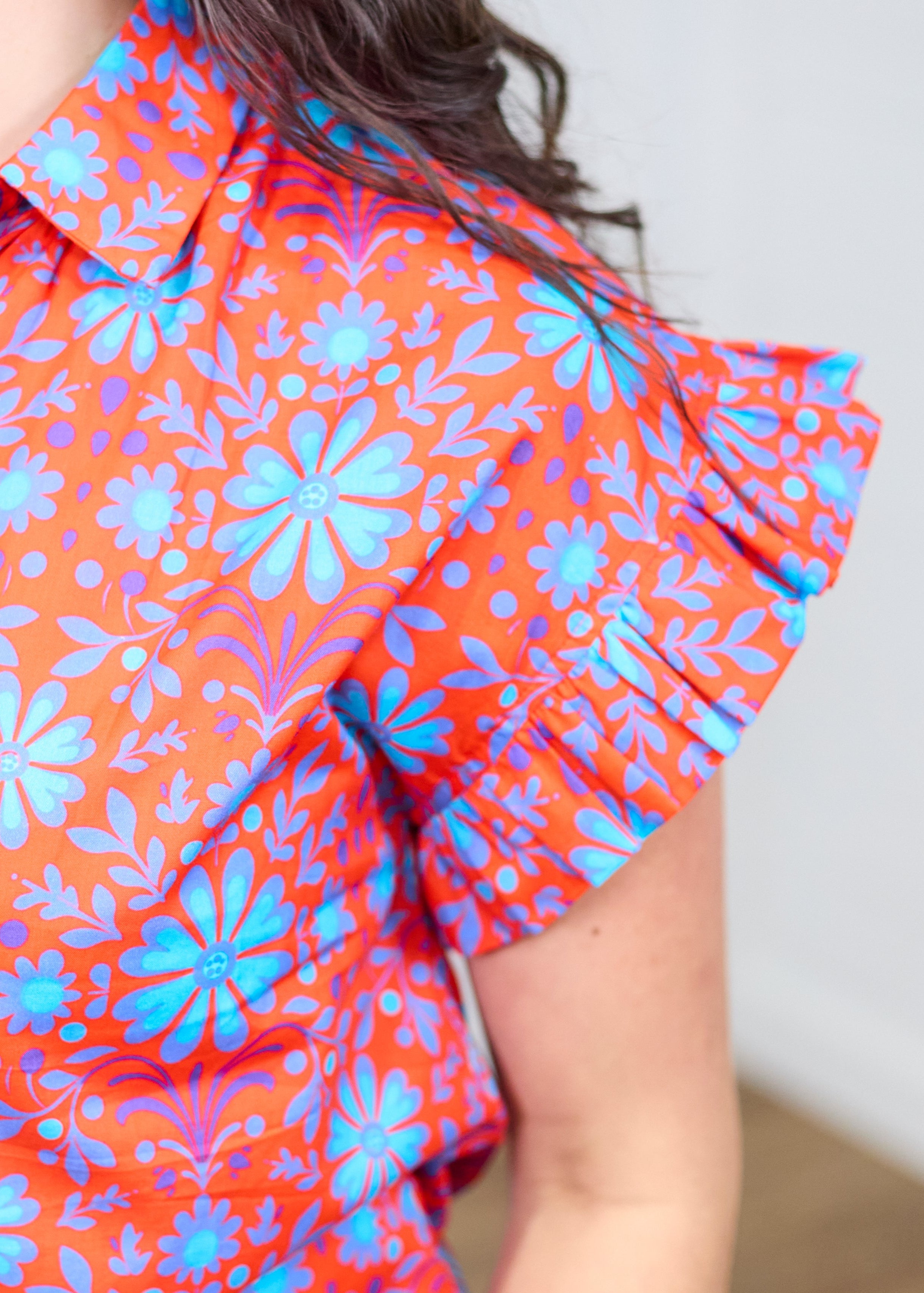Woman with dark hair standing in a front profile position wearing the Stevie Top against a white background. The focal point of this image is the deep focus on the ruffled shoulder and graphics on shirt.