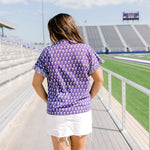 Woman with dark hair standing in back profile wearing the Stevie Top at a football field. The focal point for this image is the graphics on the back of the top as well as the striped ruffle on sleeves. The Model paired this top with a white denim skirt.