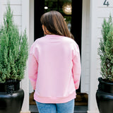 Woman with dark hair wearing a baby pink sweatshirt with embroidered Christmas motifs on front and sleeves of top. Model paired this top with light-wash denim jeans.