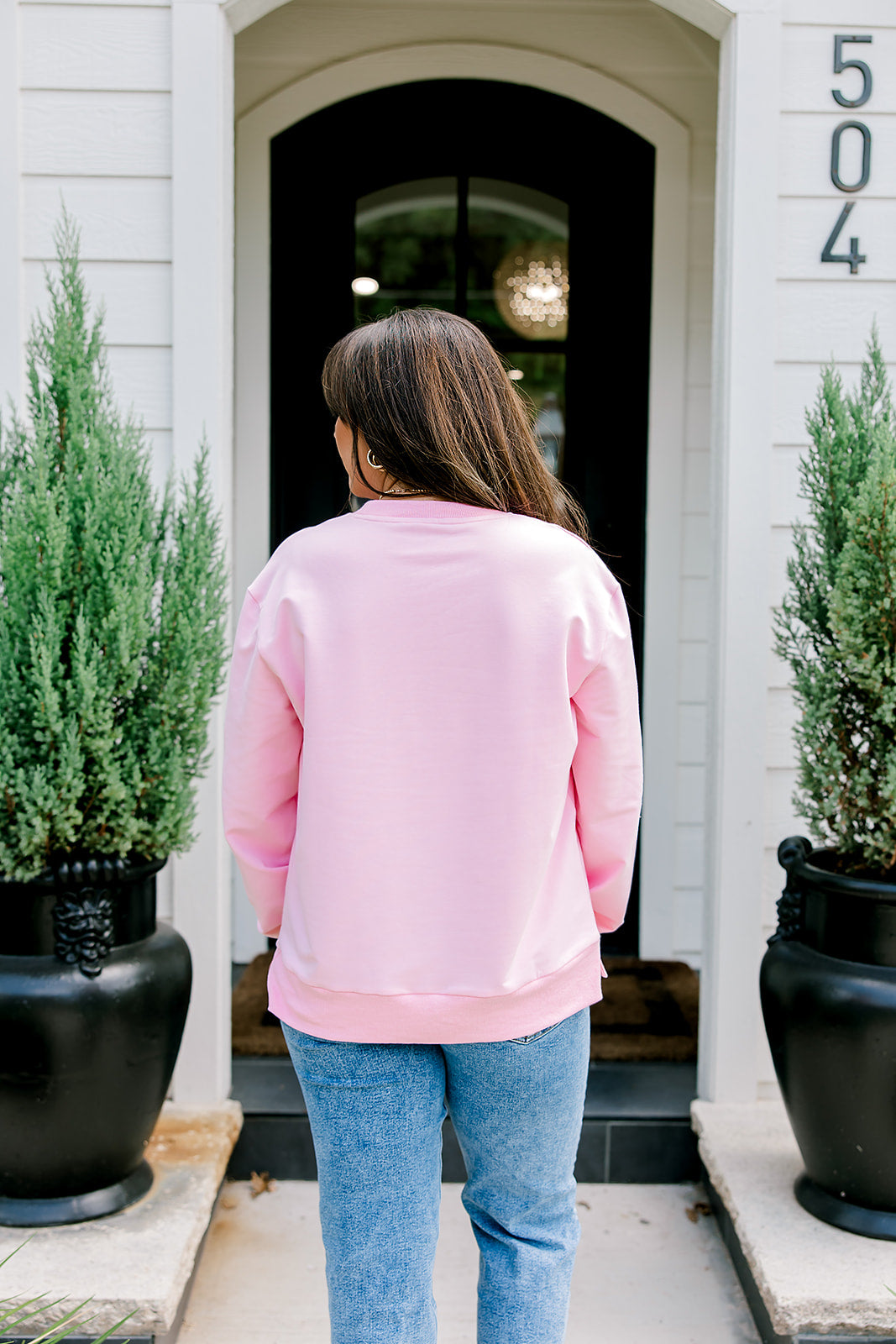 Woman with dark hair wearing a baby pink sweatshirt with embroidered Christmas motifs on front and sleeves of top. Model paired this top with light-wash denim jeans.