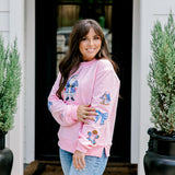Woman with dark hair wearing a baby pink sweatshirt with embroidered Christmas motifs on front and sleeves of top. Model paired this top with light-wash denim jeans.