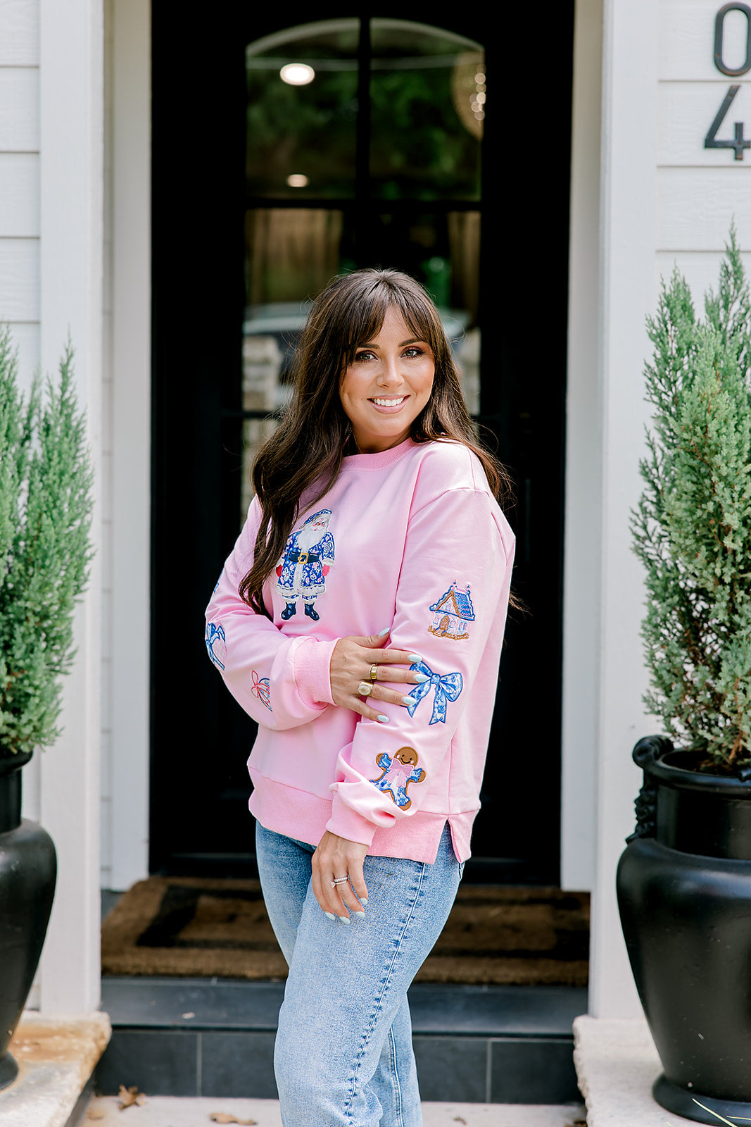 Woman with dark hair wearing a baby pink sweatshirt with embroidered Christmas motifs on front and sleeves of top. Model paired this top with light-wash denim jeans.