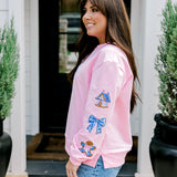 Woman with dark hair wearing a baby pink sweatshirt with embroidered Christmas motifs on front and sleeves of top. Model paired this top with light-wash denim jeans.