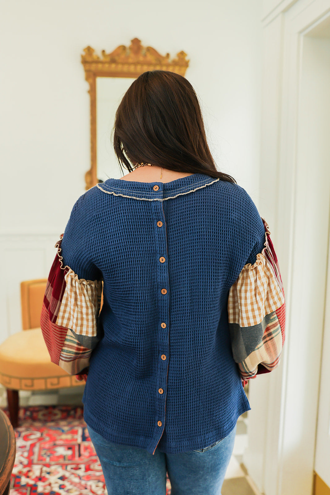Woman with dark hair standing in a back profile position wearing the Tammy Top in a lifestyle scene. The focal point of this image is the buttons down back of top as well as the patch-work ruffled sleeves. The model paired this top with mid-wash denim jeans.