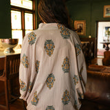 Woman with dark hair wearing a quarter sleeve poncho styled top that showcases yellow and blue floral graphics. Model paired this top with light-wash denim jeans.