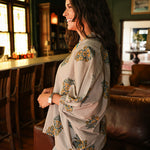 Woman with dark hair wearing a quarter sleeve poncho styled top that showcases yellow and blue floral graphics. Model paired this top with light-wash denim jeans.