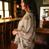 Woman with dark hair wearing a quarter sleeve poncho styled top that showcases yellow and blue floral graphics. Model paired this top with light-wash denim jeans.