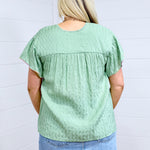 Woman with blonde hair standing in a back profile position wearing the Ada Top against a white background. The focal point of this image is the pleating on back of top as well as flowy sleeves. The model paired this top with a light-wash denim skirt. 