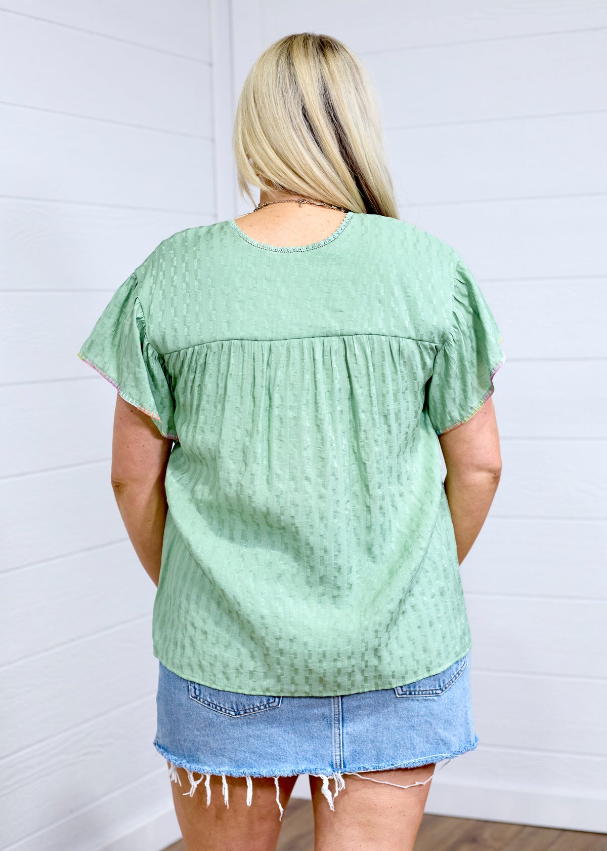 Woman with blonde hair standing in a back profile position wearing the Ada Top against a white background. The focal point of this image is the pleating on back of top as well as flowy sleeves. The model paired this top with a light-wash denim skirt. 