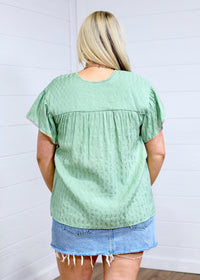 Woman with blonde hair standing in a back profile position wearing the Ada Top against a white background. The focal point of this image is the pleating on back of top as well as flowy sleeves. The model paired this top with a light-wash denim skirt. 