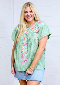 Woman with blonde hair standing in a front profile position wearing the Ada Top against a white background. The focal point of this image is the embroidery down the front, sides and chest as well as the intricate details around neckline. The model paired this top with a light-wash denim skirt. 