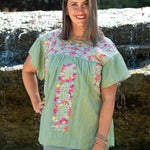 Woman with dark hair standing in a front profile position wearing the Ada Top in a lifestyle scene. The focal point of this image is the embroidery across the chest, down the front of the top as well as the neckline. The model paired this top with light-wash denim jeans.