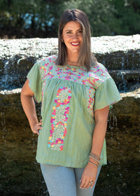 Woman with dark hair standing in a front profile position wearing the Ada Top in a lifestyle scene. The focal point of this image is the embroidery across the chest, down the front of the top as well as the neckline. The model paired this top with light-wash denim jeans.