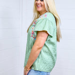 Woman with blonde hair standing in a side profile position wearing the Ada Top against a white background. The focal point of this image is the ruffle sleeve, pink buck-stitch, and embroidery on shoulders. The model paired this top with a light-wash denim skirt. 