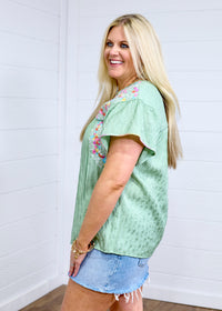 Woman with blonde hair standing in a side profile position wearing the Ada Top against a white background. The focal point of this image is the ruffle sleeve, pink buck-stitch, and embroidery on shoulders. The model paired this top with a light-wash denim skirt. 