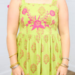 Woman with dark hair standing in a front profile position with a deep focal length towards the top embroidery on dress. This shows the floral print and the pom-poms on tied straps. 