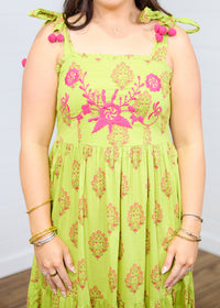 Woman with dark hair standing in a front profile position with a deep focal length towards the top embroidery on dress. This shows the floral print and the pom-poms on tied straps. 