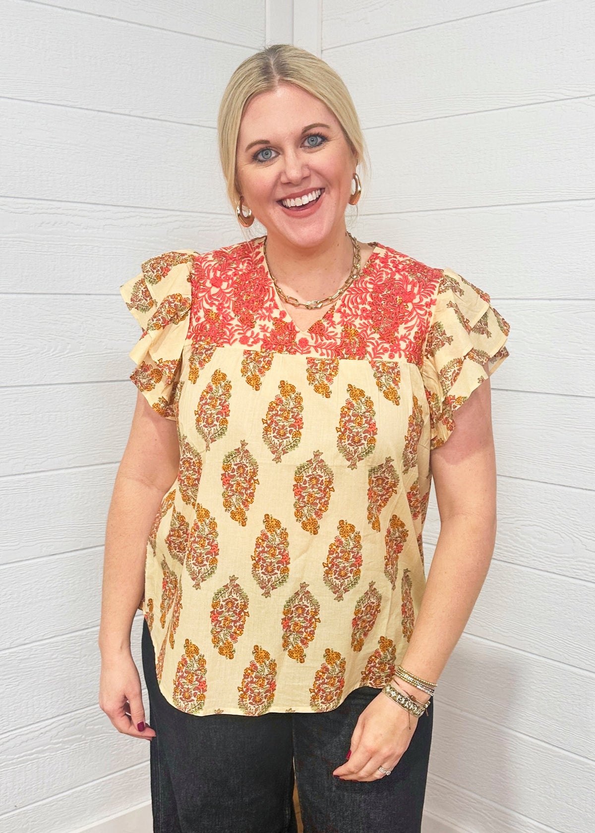 Woman with blonde hair standing in a front profile position wearing the Alura Top against a white background. The focal point of this image is the floral graphics and embroidery across chest of top. The model paired this top with dark-wash denim jeans. 