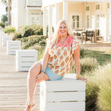Woman with blonde hair sitting in a front profile position wearing the Alura Top in a lifestyle scene. The focal point of this image is the top in its entirety; graphics, embroidery and v-neckline. The model paired this top with a light-wash denim skirt and brown sandals.