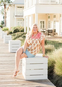 Woman with blonde hair sitting in a front profile position wearing the Alura Top in a lifestyle scene. The focal point of this image is the top in its entirety; graphics, embroidery and v-neckline. The model paired this top with a light-wash denim skirt and brown sandals.
