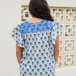 Woman with dark hair standing in a back profile position wearing the Alura Top in a lifestyle scene. The focal point of this image is the blue floral and blue embroidery. The model paired this top with light-wash denim jeans. 