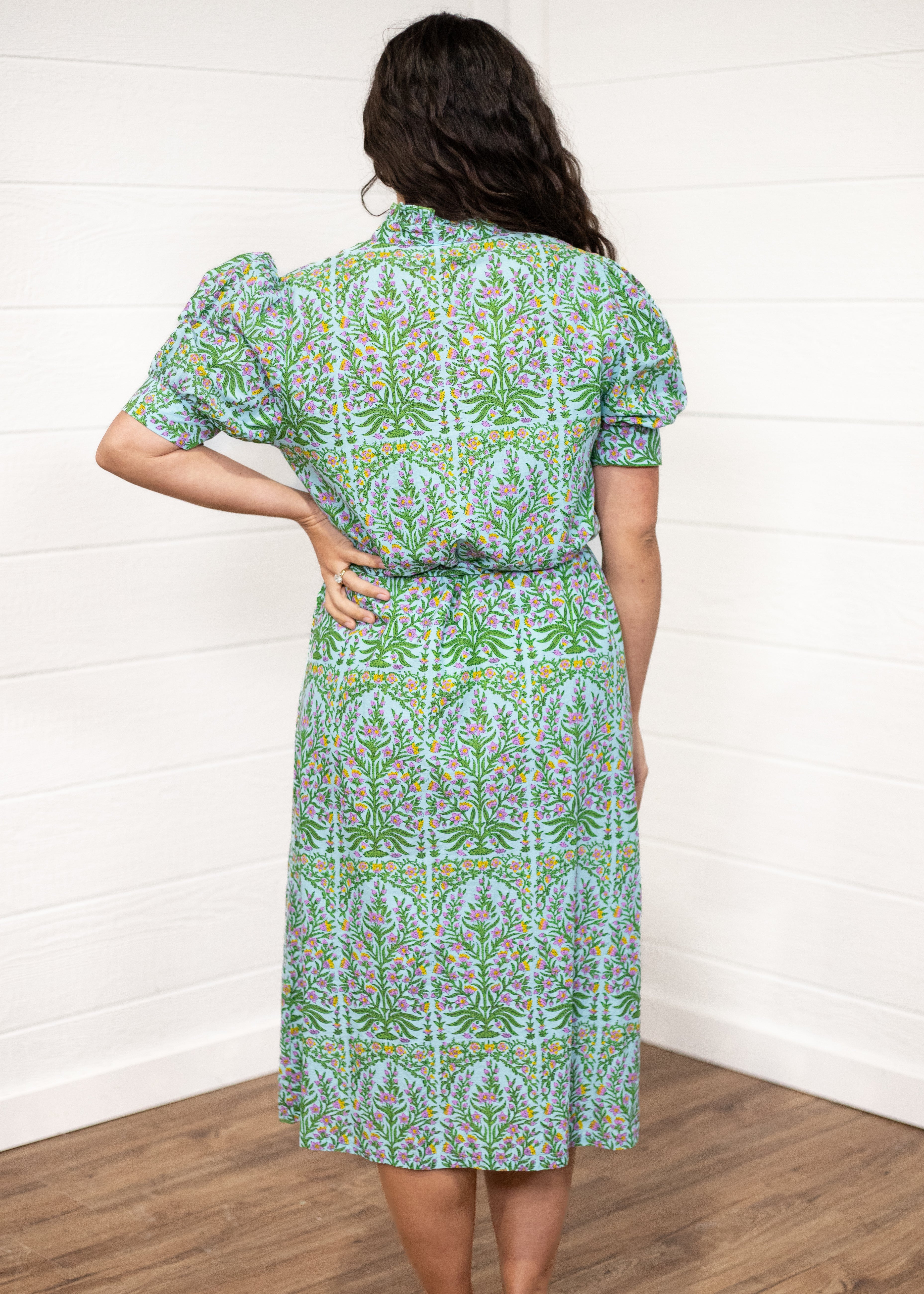 Woman with dark hair standing in a back profile position wearing the Amelia Dress against a white background. The focal point of this image is the pink, green, and orange floral graphics on the back and the puffy-cuffed sleeves.