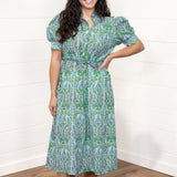Woman with dark hair standing in a front profile position wearing the Amelia Dress against a white background. The focal point of this image is the floral graphics, belt, and high collar. The model paired this dress with cream heels. 