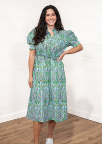 Woman with dark hair standing in a front profile position wearing the Amelia Dress against a white background. The focal point of this image is the floral graphics, belt, and high collar. The model paired this dress with cream heels. 