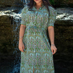 Woman with dark hair standing in a front profile position wearing the Amelia Dress in a lifestyle scene. The focal point of this image is the Amelia Dress against a water focused background. 