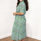 Woman with dark hair standing in a side profile position wearing the Amelia Dress against a white background. The focal point of this image is the length of dress as well as floral graphic details. The model paired this top with cream heels. 