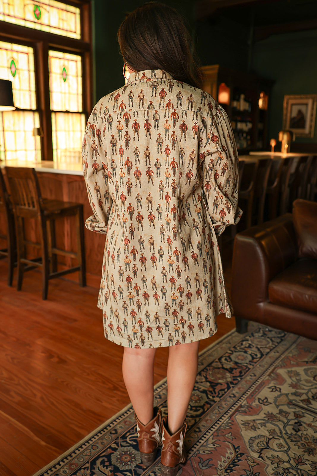 Woman with dark hair wearing a midi long-sleeve dress with cowboy graphics. Model paired this dress with brown and white booties. 