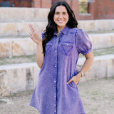 Woman with dark hair standing in a front profile position wearing the Annie Dress in a lifestyle scene. The focal point of this image is the pockets on front of dress, ruffles sleeves, pearl snaps, and length of dress. 
