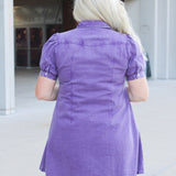 Woman with blonde hair standing in a back profile position wearing the Annie Dress in a lifestyle scene. The focal point of this image is the purple denim fabric as well as puffy sleeves. 