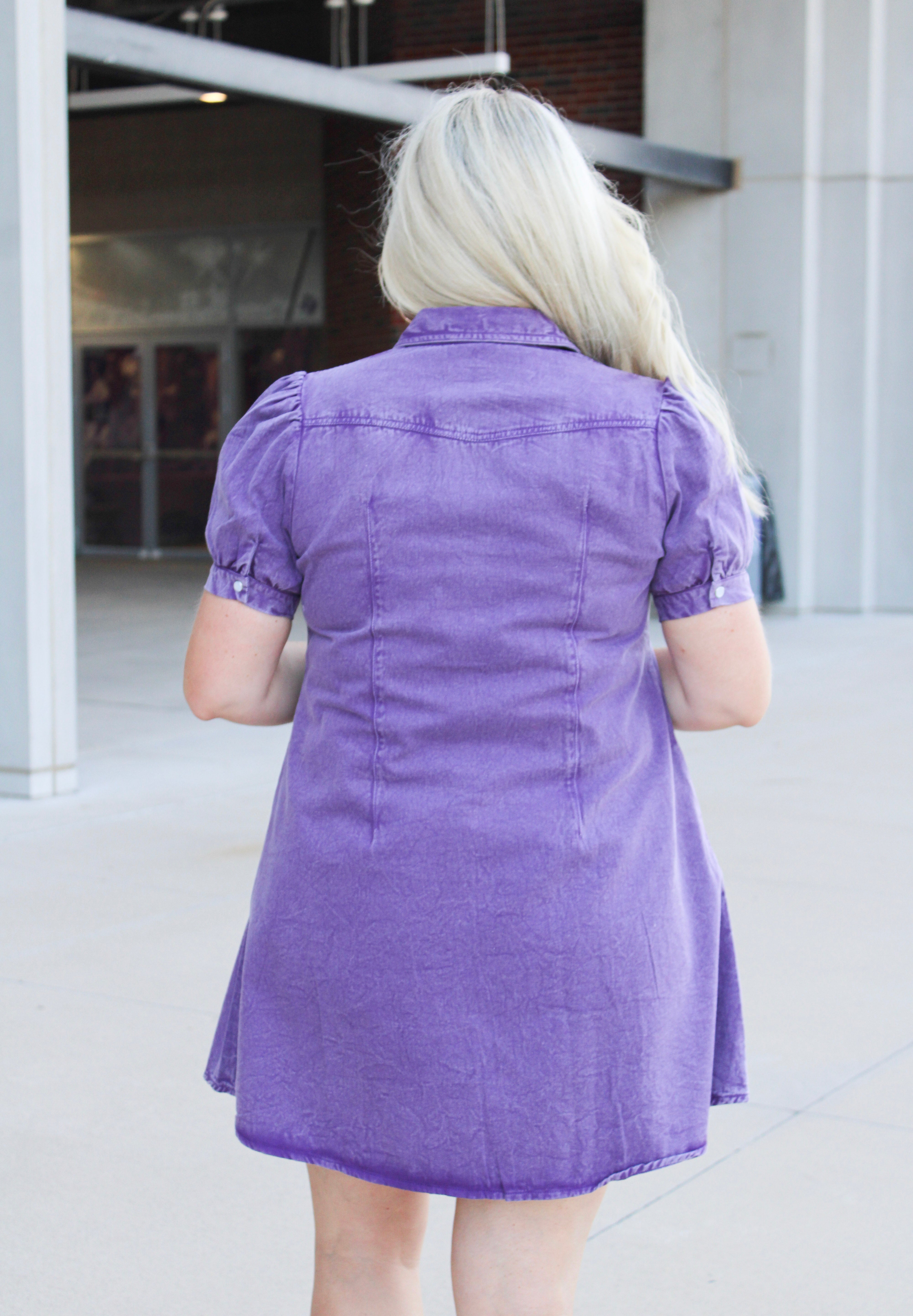 Woman with blonde hair standing in a back profile position wearing the Annie Dress in a lifestyle scene. The focal point of this image is the purple denim fabric as well as puffy sleeves. 