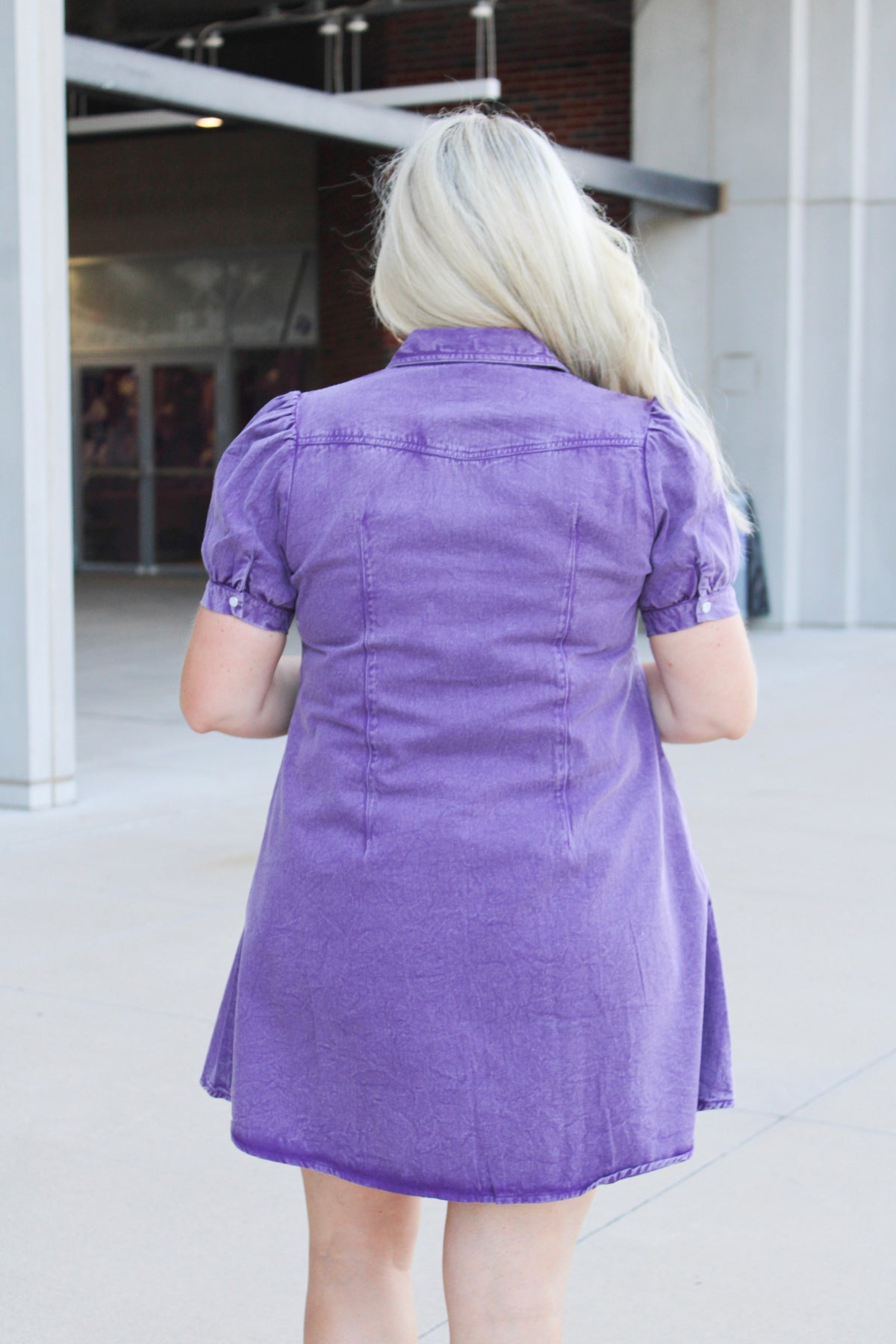 Woman with blonde hair standing in a back profile position wearing the Annie Dress in a lifestyle scene. The focal point of this image is the purple denim fabric as well as puffy sleeves. 