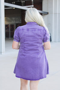 Woman with blonde hair standing in a back profile position wearing the Annie Dress in a lifestyle scene. The focal point of this image is the purple denim fabric as well as puffy sleeves. 